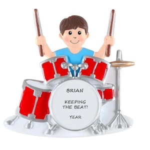 May include: A decorative ornament featuring a cartoon drummer with a blue shirt and brown hair, holding drumsticks. The red drums have silver accents. The text on the drum reads "BRIAN KEEPING THE BEAT! YEAR".