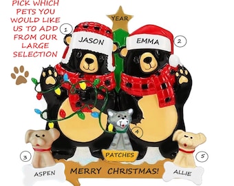 May include: A Christmas ornament featuring two black bear figures wearing red and black plaid scarves and Santa hats. The ornament includes the names "Jason" and "Emma", with attached dog and cat figures. The text "Merry Christmas!" is also present.