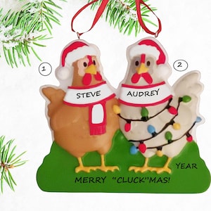 May include: A Christmas ornament featuring two cartoon chickens wearing Santa hats and scarves. One chicken is brown, the other is white, and both are decorated with Christmas lights. The ornament says "Steve" and "Audrey" on the scarves and "Merry 'Cluck'mas!" at the bottom.
