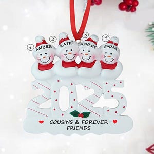 May include: A white Christmas ornament featuring four snowmen with red scarves and hats, each with a name: Amber, Katie, Ashley, and Emma. The ornament reads "2025" and "Cousins & Forever Friends."