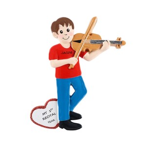 May include: A personalized Christmas ornament featuring a boy playing a violin. The ornament is red, blue, and brown. The boy is wearing a red shirt with the name "Jacob" on it. The ornament also includes a heart-shaped plaque with the text "My 1st Recital Year".