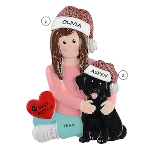May include: A Christmas ornament featuring a figure holding a black dog wearing a Santa hat. The figure wears a pink shirt and blue pants. The ornament includes the names "Olivia" and "Aspen", and the text "Best Buddy!" on a red heart.