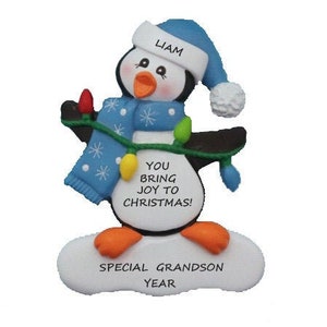 May include: Personalized Christmas ornament featuring a penguin wearing a blue hat and scarf, holding a string of lights. The ornament reads "You bring joy to Christmas!" and "Special Grandson Year".
