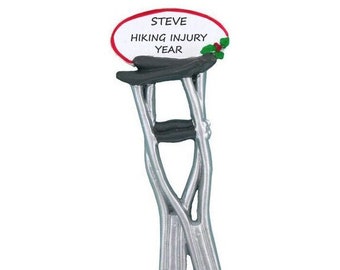 May include: A silver pair of crutches with black hand grips and black tips. A white oval ornament with red trim reads "STEVE HIKING INJURY YEAR".