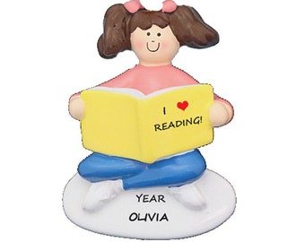 May include: A personalized ornament featuring a girl sitting with a book that says "I love reading!" The ornament is white with the words "Year Olivia" on the base.