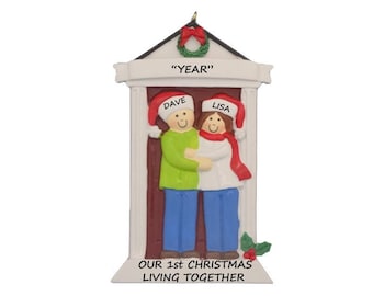 May include: A personalised Christmas bauble featuring a couple standing in a doorway. The bauble is white with a red door and a green wreath above the door. The text "YEAR" is above the wreath. The couple are wearing red Santa hats and the text "DAVE" and "LISA" is above their heads. The text "OUR 1st CHRISTMAS LIVING TOGETHER" is at the bottom of the bauble.