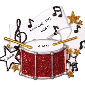 May include: Red glitter drum with a silver rim and two drumsticks. The drum has the name "Adam" written on it. A white banner with the text "Keeping the beat!" and musical notes is behind the drum. There are gold glitter stars and musical notes around the drum.