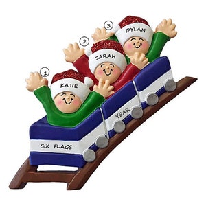 May include: A Christmas ornament depicting a family of three riding a roller coaster. The coaster is blue, white, and red with the words "Six Flags" and "Year" on it. The family members are wearing Santa hats and have names written on them: Katie, Sarah, and Dylan.