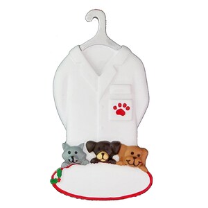 May include: White ceramic ornament shaped like a veterinarian's coat with a red paw print on the pocket. Three small ceramic animals, a grey cat and two brown dogs, are peeking out from behind the coat. The ornament has a blank oval space for personalisation.