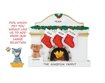 May include: A white ceramic Christmas ornament shaped like a fireplace with stockings hanging from the mantle. The stockings are red with white trim and have the names "Dad", "Mom", and "Olivia" written on them. The fireplace has a black and white firebox with a yellow flame. The ornament also includes a gray cat and a brown dog. The text "PICK WHICH PET YOU WOULD LIKE US TO ADD FROM OUR LARGE SELECTION" is written on the left side of the ornament. The text "YEAR" is written above the mantle. The text "THE KINGSTON FAMILY" is written below the fireplace.
