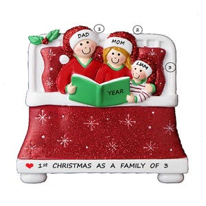 May include: A red and white Christmas ornament depicting a family of three in a bed reading a book. The ornament is decorated with white snowflakes and the text "1st Christmas as a family of 3".