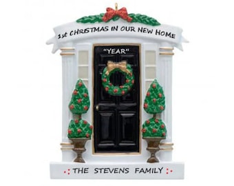 Puede incluir: Adorno navideño de cerámica blanca que representa una puerta delantera negra con una corona, dos arbustos verdes y el texto "1ª Navidad en nuestra nueva casa" y "La familia Stevens".