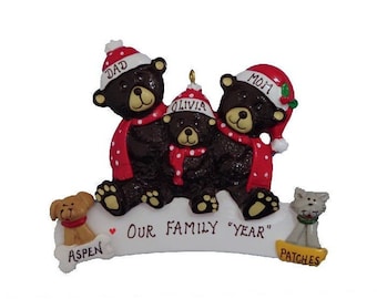 May include: A Christmas ornament featuring a family of three brown bears wearing Santa hats and scarves. The bears are sitting on a white banner that reads "Our Family "Year"". There are two pets, a brown dog named Aspen and a white cat named Patches.