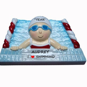 May include: A personalized Christmas ornament featuring a cartoon swimmer with a red swimsuit, blue goggles, and a white swim cap. The ornament is decorated with a blue pool and red and white lane lines. The text "Audrey I &hearts; SWIMMING!" is printed on the ornament.