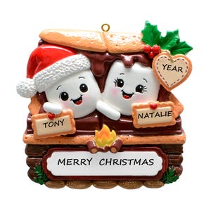 May include: A Christmas ornament featuring two marshmallow characters with brown chocolate and graham cracker details. One marshmallow wears a red Santa hat. The ornament includes signs labeled TONY, NATALIE, and YEAR, with a banner reading MERRY CHRISTMAS.