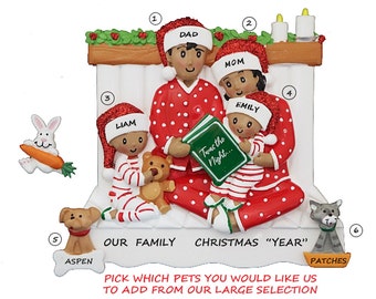 Puede incluir: Un adorno navideño personalizado que representa a una familia de cuatro personas en pijama rojo y blanco, leyendo un libro junto a una chimenea. El adorno incluye los nombres "Papá", "Mamá", "Liam" y "Emily". También se incluye el texto "Nuestra familia Navidad "Año"". El adorno está decorado con acebo y velas.