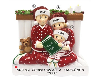 May include: A personalized Christmas ornament featuring a family of three in red and white pajamas, sitting by a fireplace with a book titled "Twas the Night..." The ornament is decorated with a green garland and candles. The text "OUR 1st CHRISTMAS AS A FAMILY OF 3 "YEAR" is printed on the bottom.