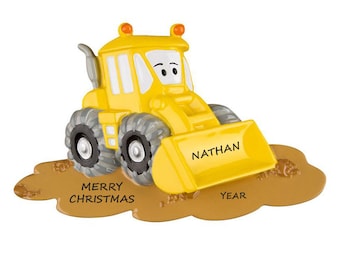 Pode incluir: Ornamento de veículo de construção de brinquedo amarelo com um rosto sorridente. O veículo está cavando na terra marrom com as palavras "Feliz Natal" e "Ano" na terra. A lâmina do veículo tem o nome "Nathan" escrito nela.