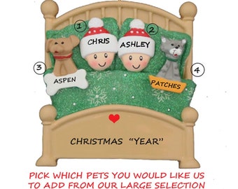 May include: A personalized Christmas ornament featuring a bed with two people and two pets. The bed is brown with a green blanket and white snowflakes. The people are wearing red Santa hats and have names "Chris" and "Ashley". The pets are a dog named "Aspen" and a cat named "Patches". The ornament says "Christmas "Year"".