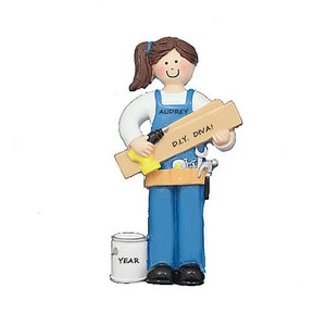 May include: A personalized Christmas ornament featuring a woman wearing blue overalls and a tool belt. She is holding a piece of wood and a drill. The ornament also includes a paint can and the text "Audrey D.I.Y. DIVA!" and "YEAR".