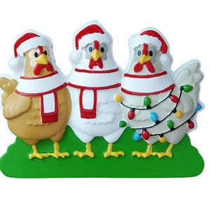 May include: A decorative Christmas ornament featuring three chickens wearing Santa hats and scarves. The chickens are in shades of brown and white, with red and white accents. One chicken is adorned with colorful Christmas lights, all standing on a green base.