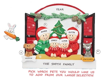 May include: A personalized Christmas ornament featuring a red door with a family of four standing in front of a Christmas tree. The ornament includes the names "Mark", "Katie", "Emily", and "Liam", and the text "The Smith Family". There are two pets, a dog named "Aspen" and a cat named "Patches".