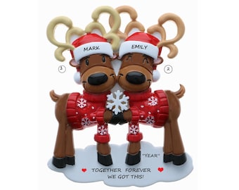 May include: Personalized Christmas ornament featuring two reindeer wearing red sweaters with snowflakes, holding a snowflake together. The reindeer are wearing Santa hats with the names "Mark" and "Emily" written on them. The ornament also includes the text "Together Forever We Got This!" and "Year".