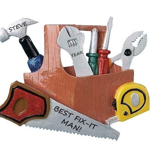 May include: A brown wooden toolbox ornament with a saw, hammer, wrench, pliers, and a tape measure. The toolbox has the text "BEST FIX-IT MAN!" and "STEVE YEAR" on it.