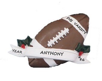 May include: A brown and white football ornament with a white banner that reads "YEAR ANTHONY #86" and a green holly sprig on each side. The football has the text "FHS VARSITY" on the side.
