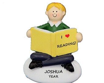 May include: A personalized ornament featuring a boy sitting with a book that says "I love reading!" The ornament is white with the name "Joshua" and the word "Year" written on it.