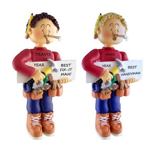 May include: Two personalized Christmas ornaments depicting a cartoon handyman holding a sign and tools. The ornaments are red, blue, and brown. The sign on the left ornament says "Best Fix-It Man!" and the sign on the right ornament says "Best Handyman".