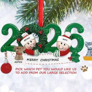 May include: A personalized Christmas ornament with the year 2023 in green. It features two figures wearing Santa hats, a dog, a cat, and the text "MERRY CHRISTMAS". The ornament includes the names "BRIAN" and "AMY".