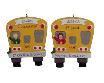 May include: Two yellow school bus ornaments with a boy and girl in the windows. The bus on the left says "Connor Kindergarten 1st Bus Ride to School" and the bus on the right says "Jessica 1st Grade Sycamore Elementary".