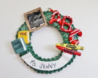 May include: Green wreath ornament with a chalkboard that says "A+ Teacher", two books that say "Year" and "From Carter", red and white ABC letters, two red and yellow crayons that say "Crayon", and a white banner that says "Ms. Jenny".