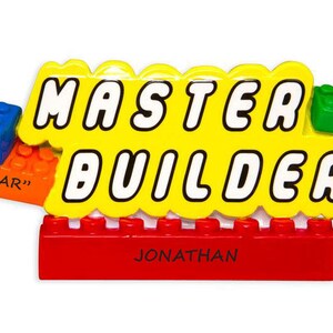 May include: A colorful ornament in the style of toy building blocks. The yellow centerpiece reads "MASTER BUILDER" in white letters. Additional blocks are blue, orange, and green. The name "JONATHAN" is printed on a red block.