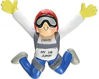 May include: A personalized skydiving ornament featuring a cartoon figure in a skydiving suit with a red helmet, goggles, and a white shirt. The figure is wearing blue pants and has a gray parachute harness with the text "DAVID MY 1ST JUMP!" and "YEAR" written on it.