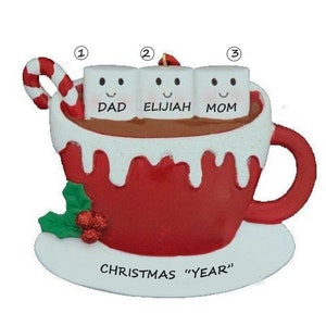 Puede incluir: Un adorno navide&ntilde;o rojo y blanco con forma de taza de chocolate caliente con tres malvaviscos. Los malvaviscos est&aacute;n etiquetados como "Pap&aacute;", "El&iacute;as" y "Mam&aacute;". El adorno tambi&eacute;n tiene un bast&oacute;n de caramelo y hojas de acebo.