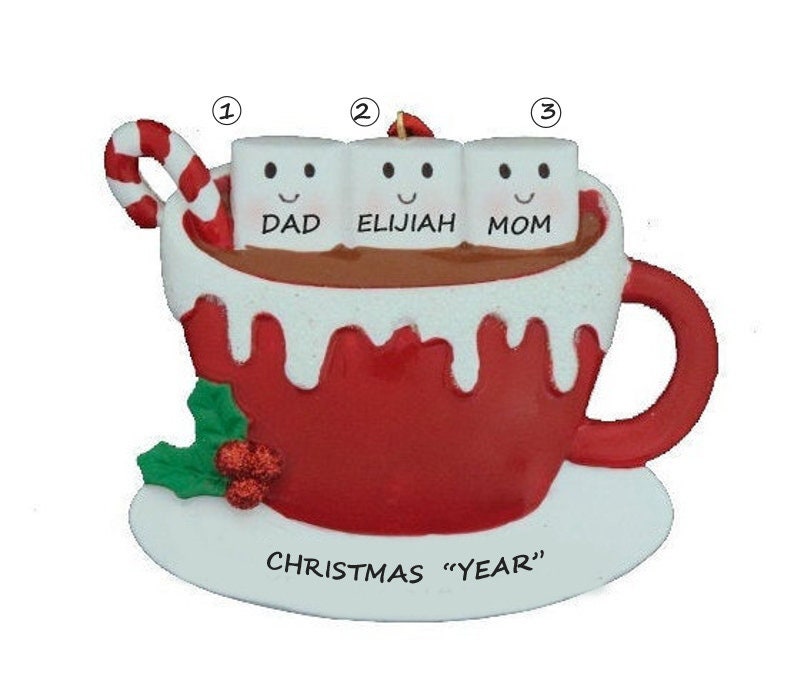 Puede incluir: Un adorno navide&ntilde;o rojo y blanco con forma de taza de chocolate caliente con tres malvaviscos. Los malvaviscos est&aacute;n etiquetados como "Pap&aacute;", "El&iacute;as" y "Mam&aacute;". El adorno tambi&eacute;n tiene un bast&oacute;n de caramelo y hojas de acebo.