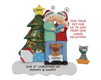 May include: Personalized Christmas ornament featuring a couple holding a baby, a dog, and a Christmas tree. The ornament says "Our 1st Christmas as Mommy & Daddy" and includes the names "Steve", "Kelly", and "Noah".