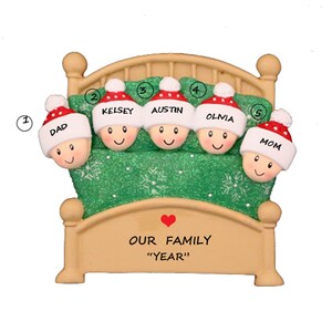 May include: A Christmas ornament depicting a family of five sleeping in a bed. The bed is brown with a green blanket and white snowflakes. The family members are wearing red and white Santa hats. The ornament has the text "Our Family "Year"" on the bottom.