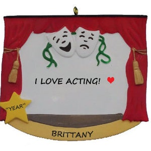 May include: A red and gold Christmas ornament with a stage curtain, two theater masks, and a blank space for a personalized message. The ornament says "I love acting!" and has a star with the word "YEAR" on it. The bottom of the ornament says "BRITTANY".