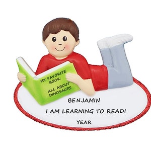 May include: A personalized Christmas ornament featuring a boy reading a book with the text "My Favorite Book: All About Dinosaurs". The ornament also includes the name "Benjamin" and the text "I am learning to read!" with a space for the year.