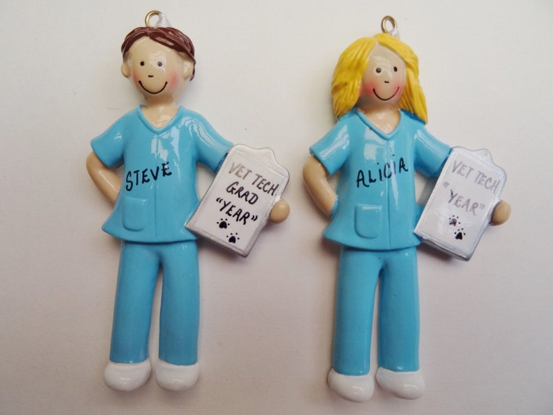 May include: Two personalized ornaments of a male and female vet tech wearing blue scrubs and holding a clipboard with the text "Vet Tech Grad "Year"". The male ornament has the name "Steve" and the female ornament has the name "Alicia".