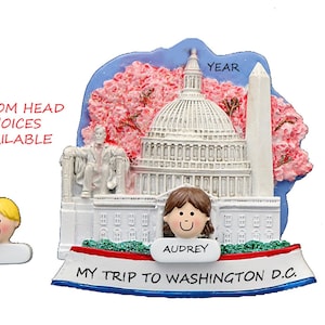 May include: White ceramic ornament featuring a depiction of the US Capitol Building with cherry blossoms, the Washington Monument, and a figure of Abraham Lincoln. The ornament has a red, white, and blue ribbon with the text "MY TRIP TO WASHINGTON D.C." and a blank space for a year. A personalized head can be added to the ornament. The text "AUDREY" is on the ornament.