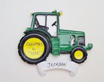 Peut inclure: Ornement de Noël en forme de tracteur vert avec une roue jaune et une bannière blanche qui dit "Jackson". La roue porte les mots "Christmas" et "Year" écrits dessus.