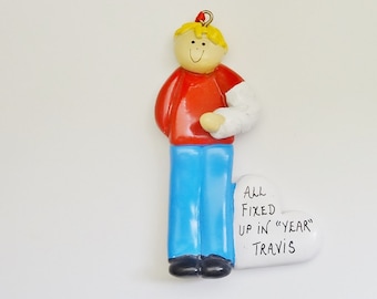 May include: A personalized Christmas ornament featuring a boy with blonde hair wearing a red shirt, blue pants, and a white cast on his arm. The ornament includes the text "All Fixed Up in "YEAR" Travis".