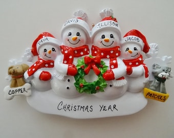 May include: A white snowman family Christmas ornament with red and white polka dot scarves and hats. The ornament includes the names "David", "Allison", "Maya", and "Jacob" and the text "Christmas Year". Two pets, a dog named "Copper" and a cat named "Patches", are included on either side of the family.