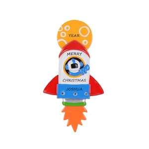 May include: A white rocket ship Christmas ornament with a red and white astronaut inside. The rocket has a red and white flame coming out of the bottom. The ornament has the words "Merry Christmas" and "Joshua" on it. There is a yellow circle with the word "Year" on it above the rocket.