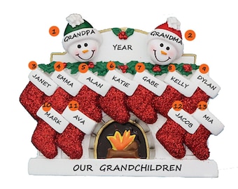 May include: A white ceramic Christmas ornament depicting a fireplace with stockings hanging from it. The stockings are red with white trim and have names written on them. The fireplace has a small fire burning in it. The ornament also features two snowmen with the words "Grandpa" and "Grandma" written on their hats. The ornament says "Our Grandchildren" at the bottom.