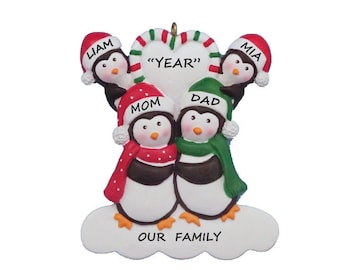 Puede incluir: Un adorno navideño personalizado con una familia de cinco pingüinos. Los pingüinos llevan gorros y bufandas rojas y verdes. El adorno está decorado con un corazón de caramelo y el texto "Nuestra familia".